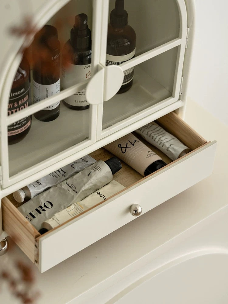 An open cream cabinet drawer holds skincare tubes beneath glass doors, with a silver ball knob at the front.