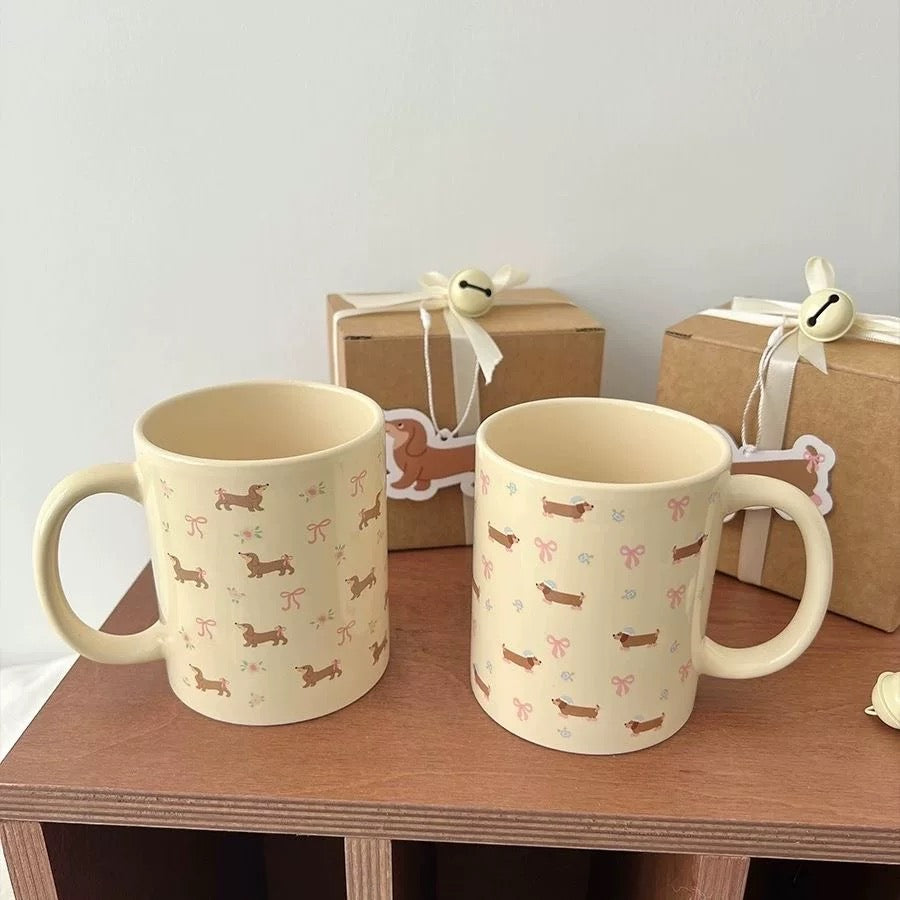 Two cream-colored ceramic mugs with dachshund and bow patterns, displayed on a wooden shelf with gift boxes in the background.