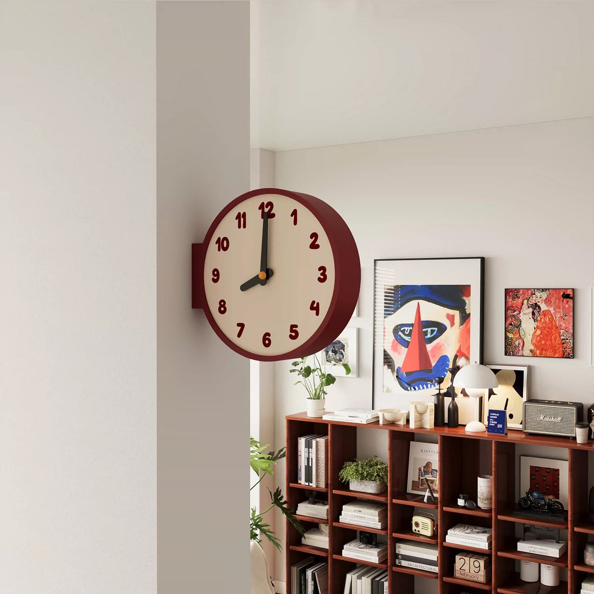 A round wall clock with a cream face, dark red outer rim, and dark red numerals, mounted on a white wall in a modern room with a wooden bookshelf and framed art below.