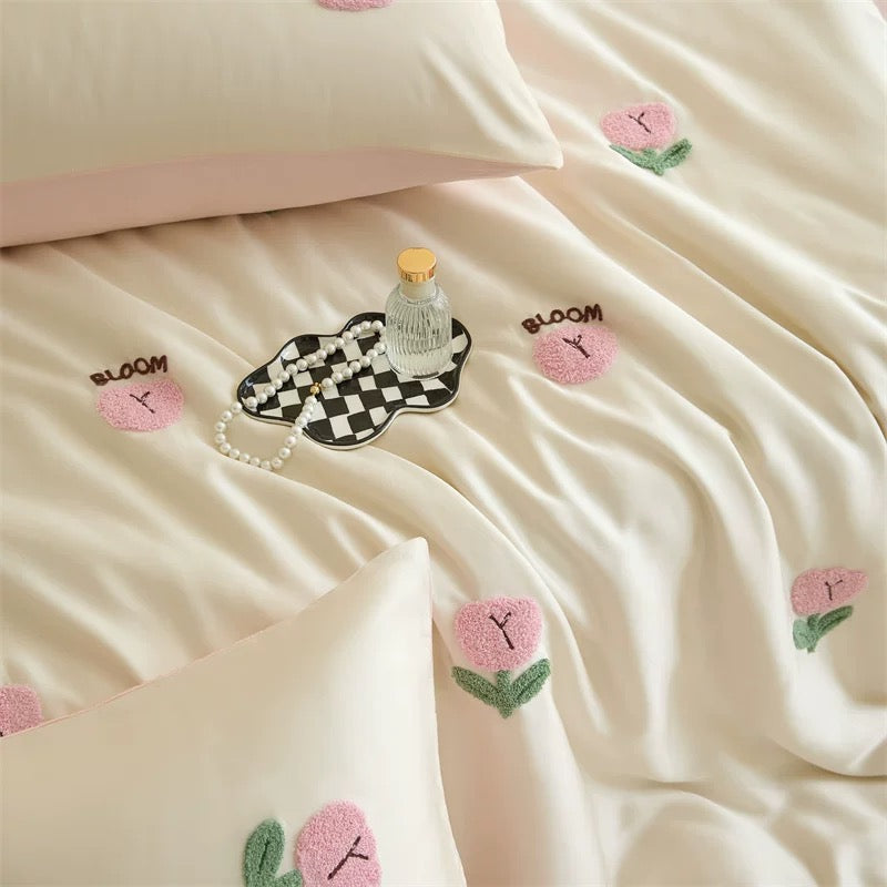 Cream-colored bedding set with embroidered pink tulips and the word 'BLOOM' on the duvet and pillowcases, styled with a small decorative tray and bottle.