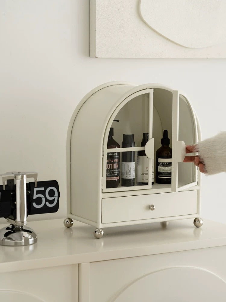 A cream makeup cabinet stands on a vanity, with bottles visible behind its doors and a drawer below.