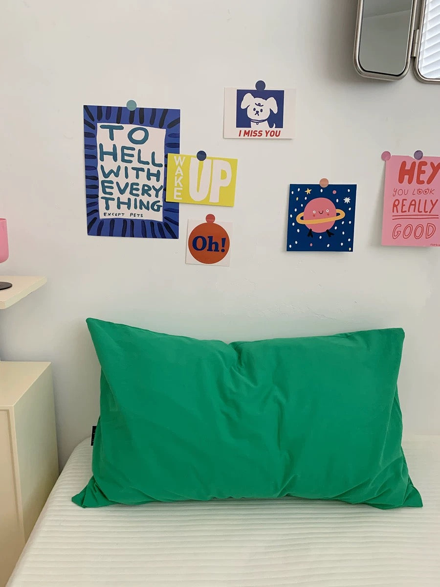A bright green rectangular pillowcase on a white bed in a bedroom, with colorful art prints pinned to a white wall above.