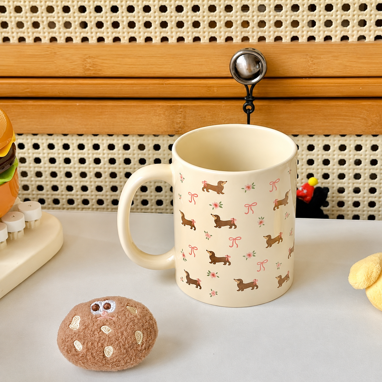A cream-colored ceramic mug with a pattern of brown dachshund dogs and pink bows, displayed on a white tabletop next to a small brown plush toy.