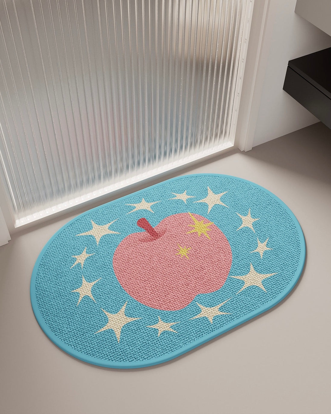 Two oval woven floor mats with apple and star designs, shown in a bathroom or entryway setting. The top mat is light blue with a pink apple and cream stars, and the bottom mat is off-white with a blue apple and pink stars.