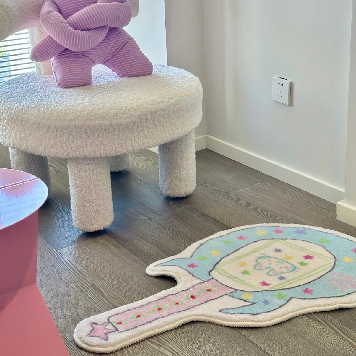 A pastel unicorn-shaped rug with a starry border and a pink horn lies on a gray wood floor in a child's room, next to a white fuzzy stool with a purple plush toy on it.