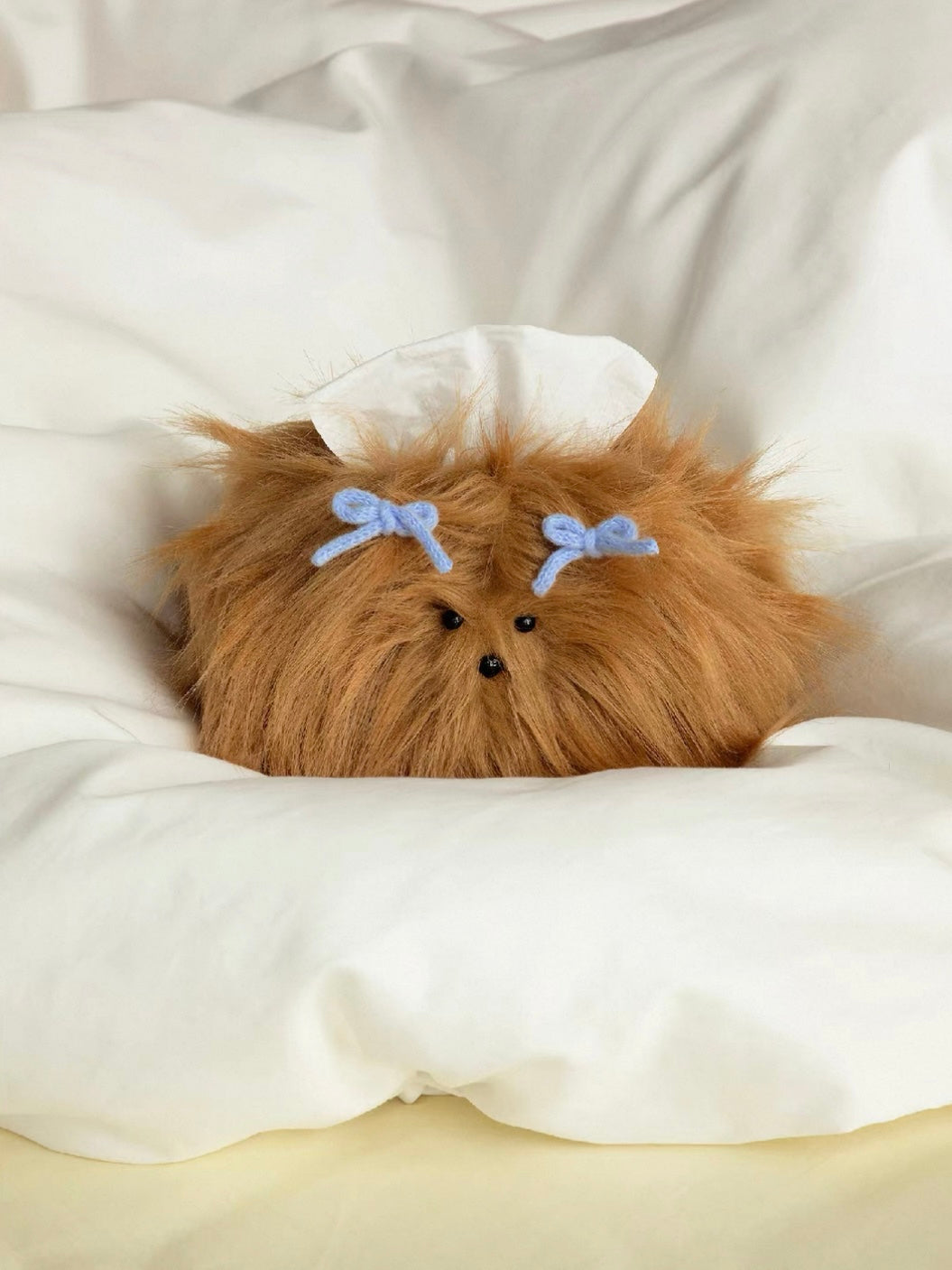 A cute tissue box cover in Caramel sits against white pillows, with blue bows above its small black eyes.