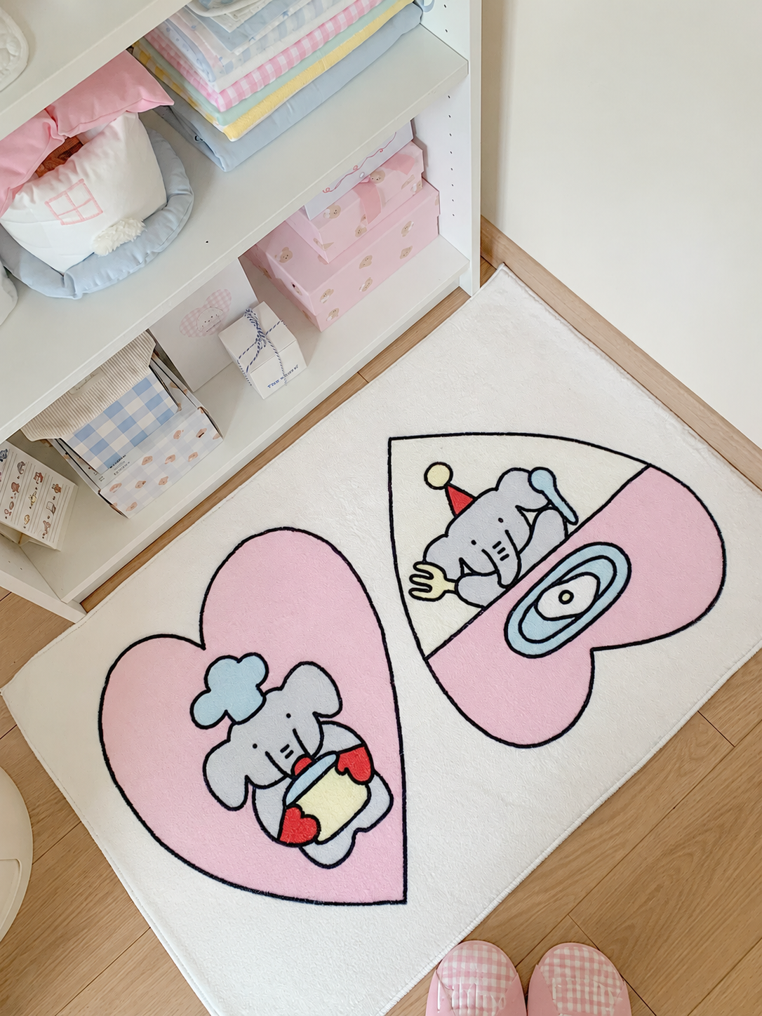A cutecore rug with two pink hearts and cooking elephants lies beside a white shelf on a cream rectangular base.