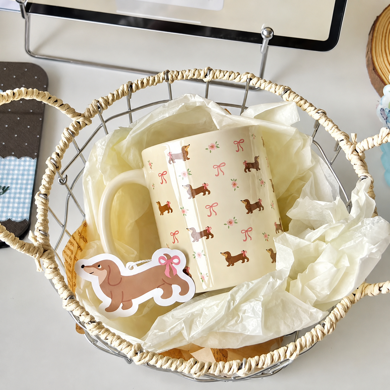 A cream-colored ceramic mug with a repeating pattern of brown dachshunds wearing pink bows, nestled inside a woven wire basket lined with white tissue paper. A matching dachshund sticker is placed on the front of the mug.
