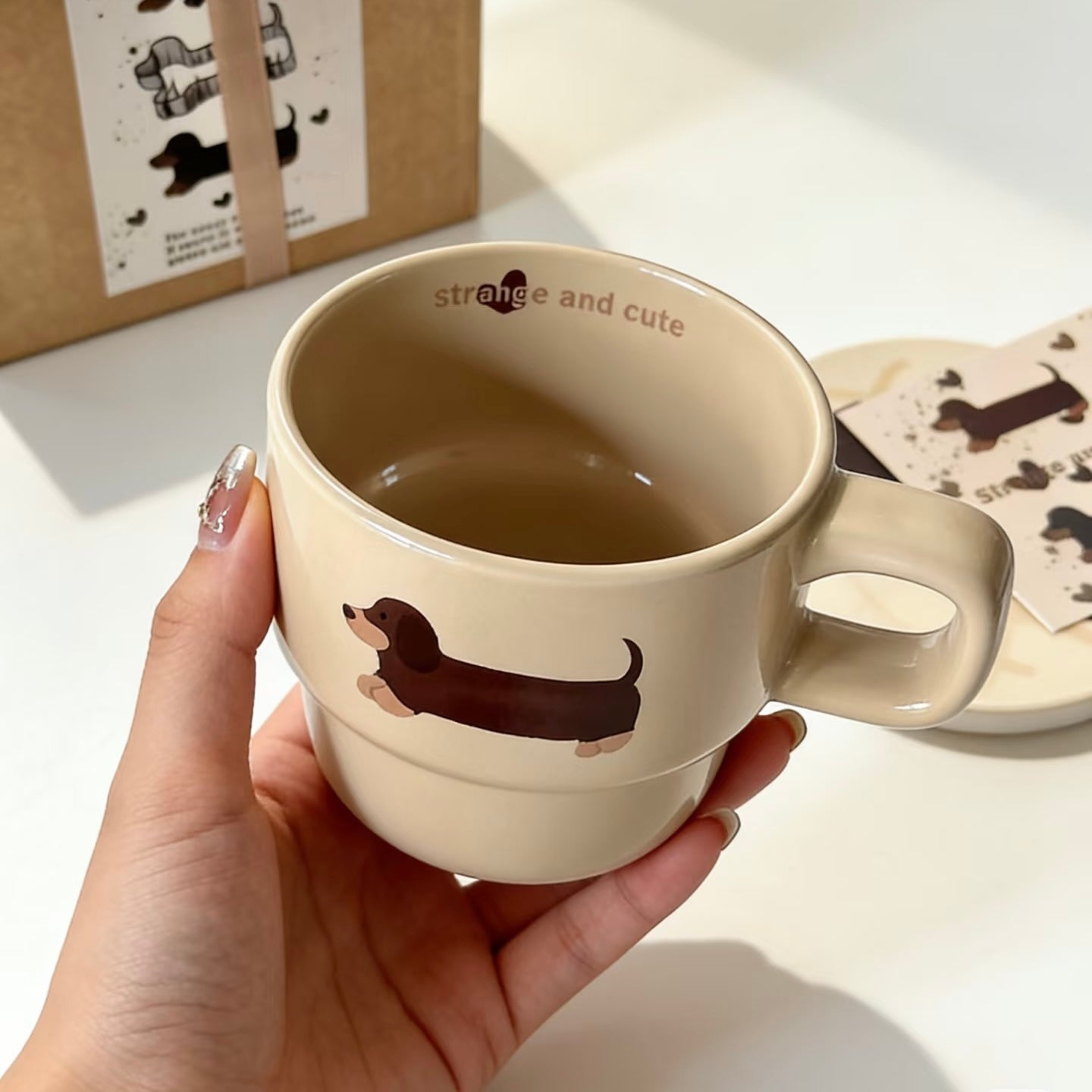 A hand holds a beige ceramic mug with a dachshund illustration and the words 'strange and cute' printed inside. A kraft paper gift box and a small card with dachshund illustrations are visible in the background on a white surface.
