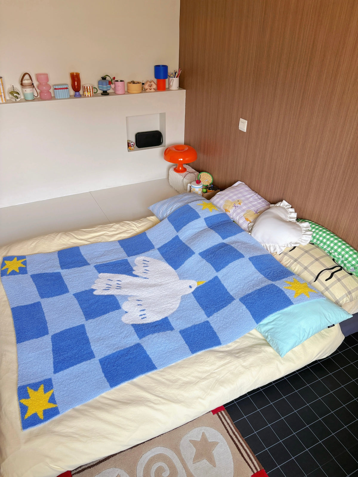 A cozy bedroom scene showing a blue checkered blanket with a white dove in the center and yellow stars in the corners, spread across a bed with cream bedding and decorative pillows.