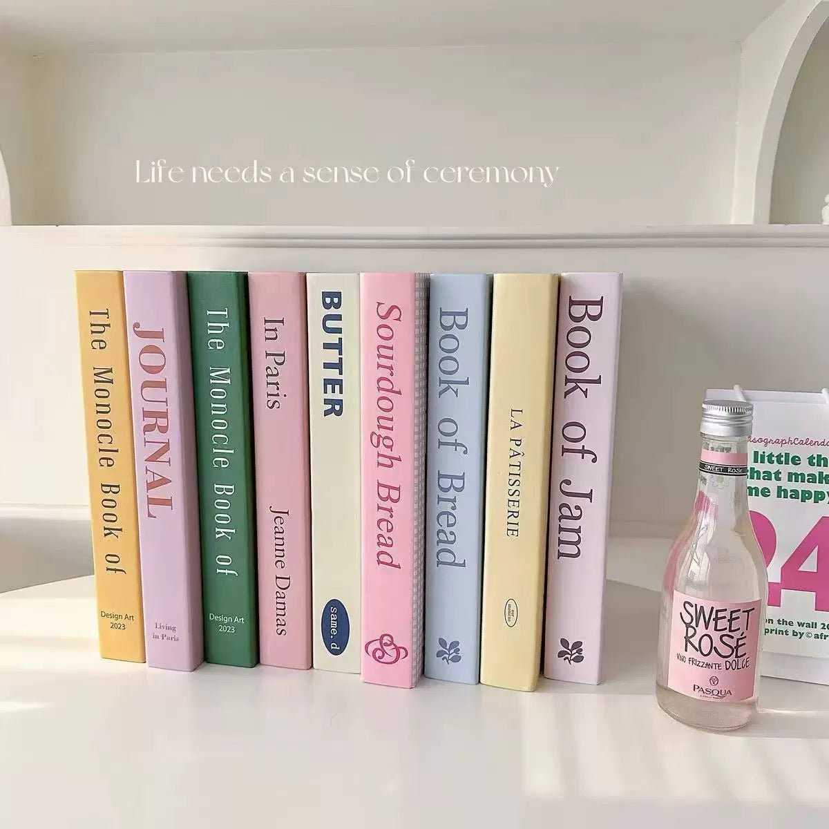 Row of books with colorful spines on a white surface, accompanied by a bottle labeled 'Sweet Rose'.
