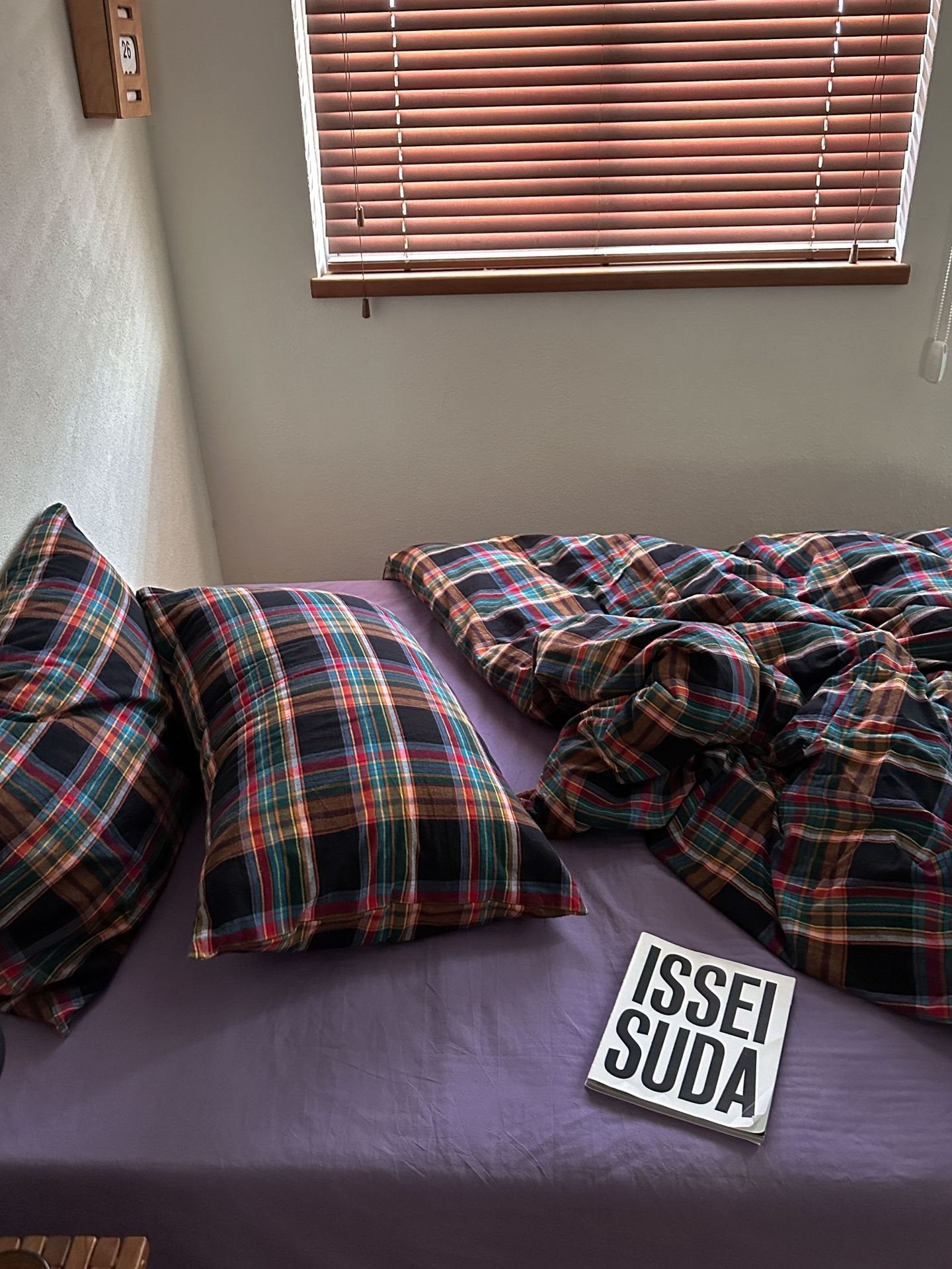 A bed with a purple sheet and matching plaid bedding in black, red, green, and blue, with a book titled 'ISSEI SUDA' on top, set in a bedroom with a window and blinds.