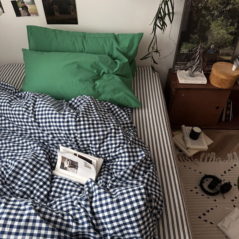 A styled bed in a bedroom setting featuring a navy and white gingham checkered duvet cover and matching pillowcases, accented by solid green pillows, with a wooden bedside table and room decor visible.