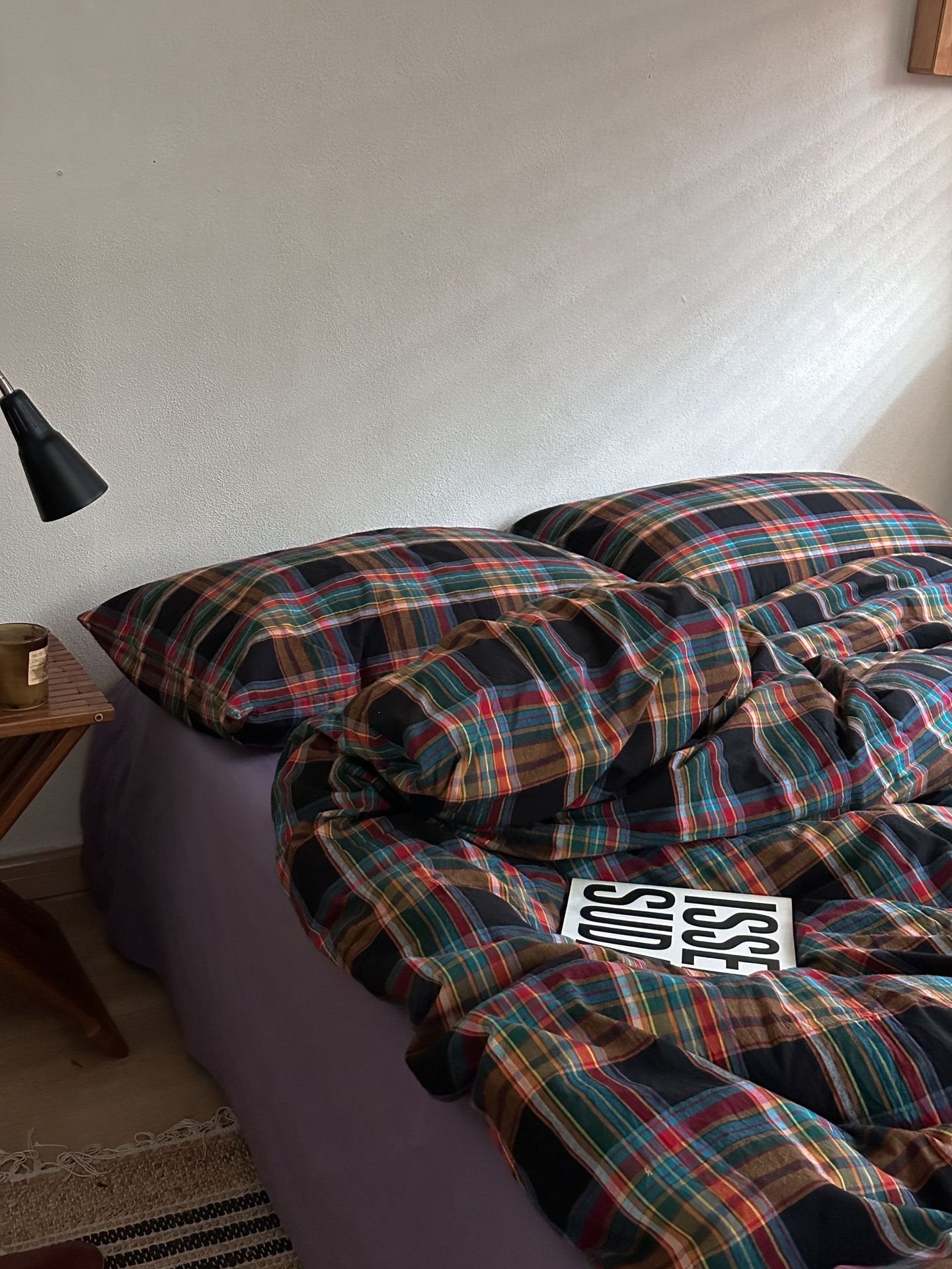 A bed in a bedroom setting with a dark plaid comforter and matching pillowcases, featuring a purple bed frame underneath. A black desk lamp is visible on the left, and a small card with the text 'MRS.