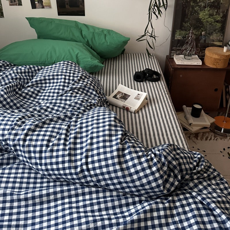A bed in a real bedroom setting with a blue and white gingham check duvet cover and matching striped bedding, plus green pillows. A wooden bedside table, books, a plant, and wall decor are visible around it.