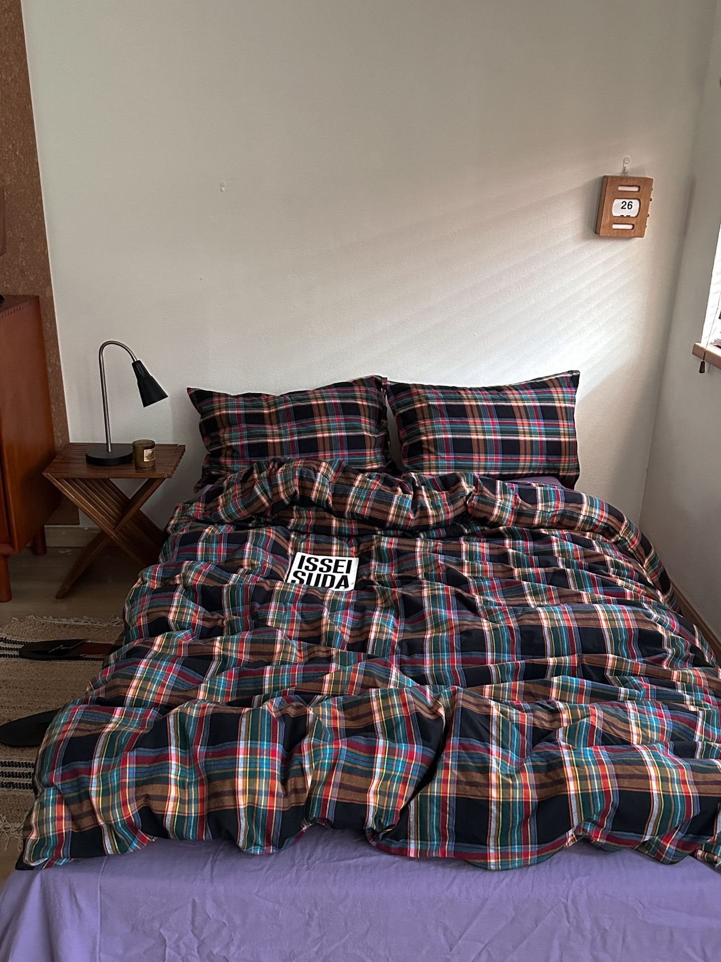 A bed dressed in a multicolor plaid comforter and matching pillowcases, with a purple sheet underneath, in a bedroom setting.