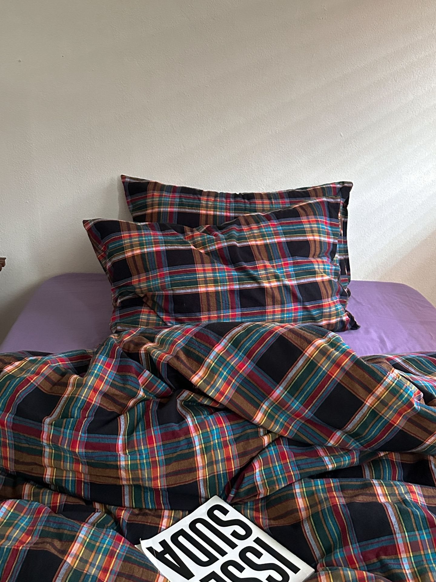 A bed dressed in a multicolor plaid bedding set with matching pillowcases, featuring a black base with red, green, blue, and yellow stripes. A purple sheet is visible underneath, and a white book with black text is placed on the bed.
