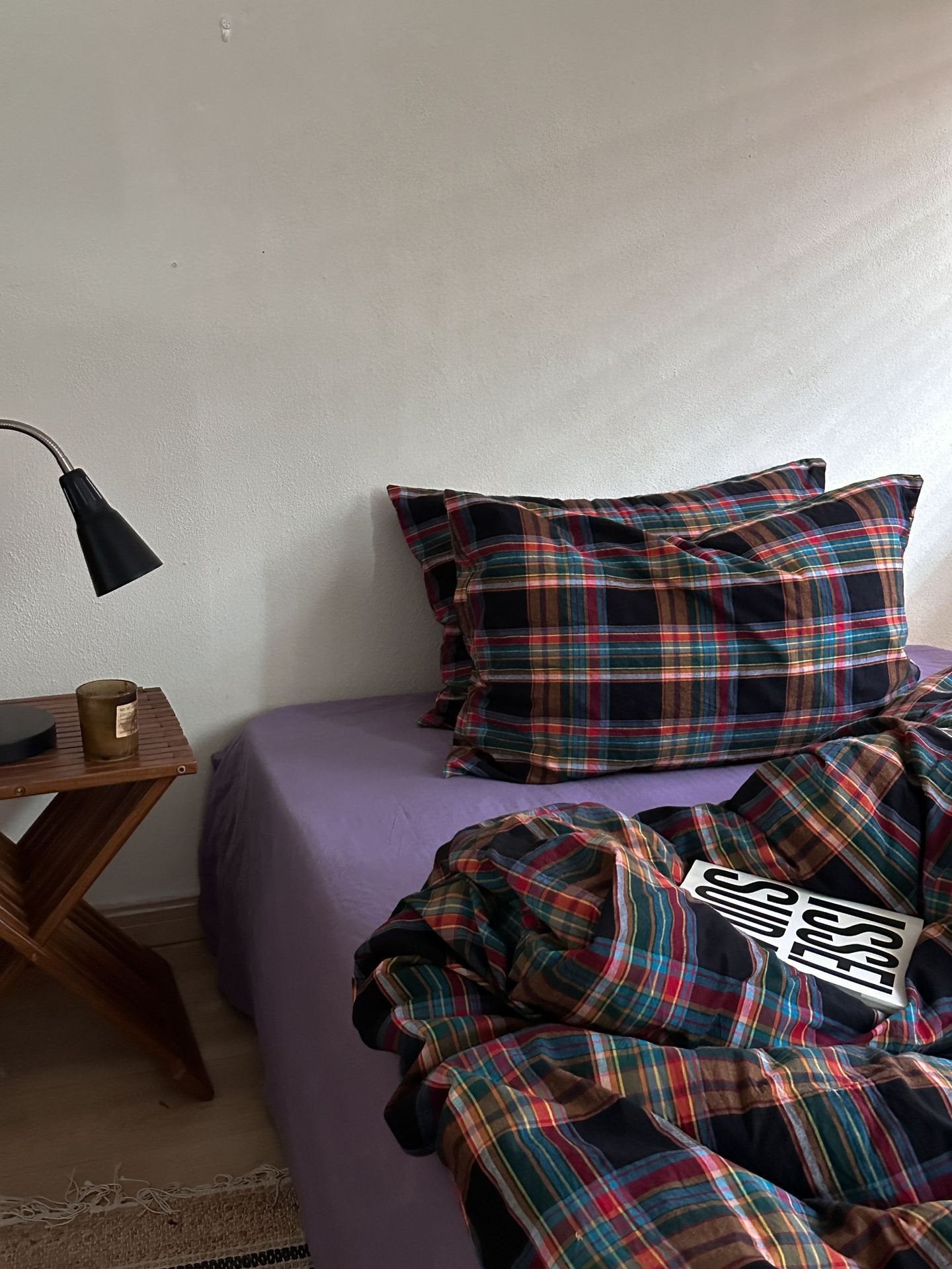 A bed in a bedroom setting featuring a purple sheet and matching plaid pillowcases, with an unmade portion of the plaid comforter in the foreground. A wooden side table with a black lamp sits to the left against an off-white wall.
