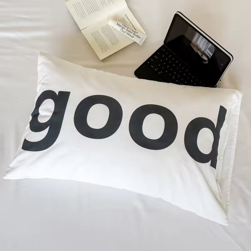 A white rectangular pillowcase with the word 'good' printed in large black lowercase letters, styled on a white bed with an open book and a black laptop nearby.