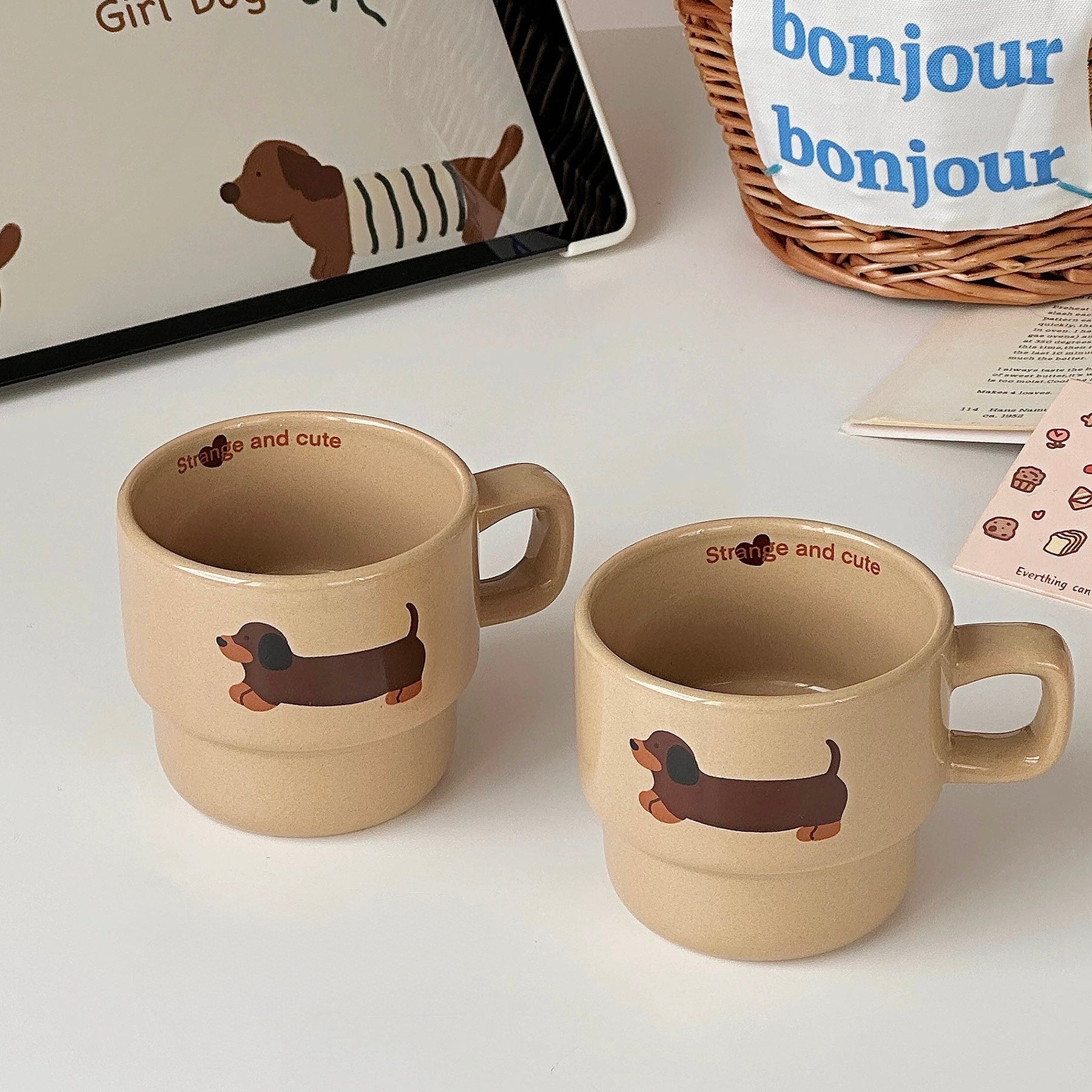 Two beige ceramic mugs with dachshund illustrations, displayed on a white surface with decorative props including a tablet, a basket, and cards.