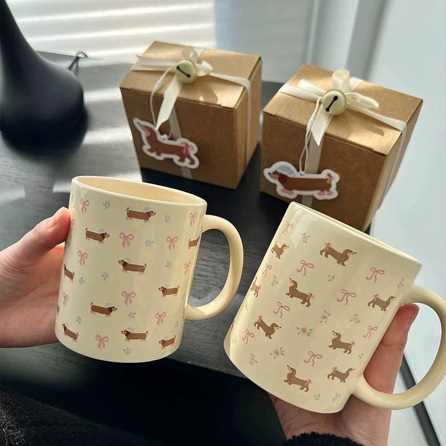 Two mugs with dachshund pattern held by a person, with two gift boxes in the background.