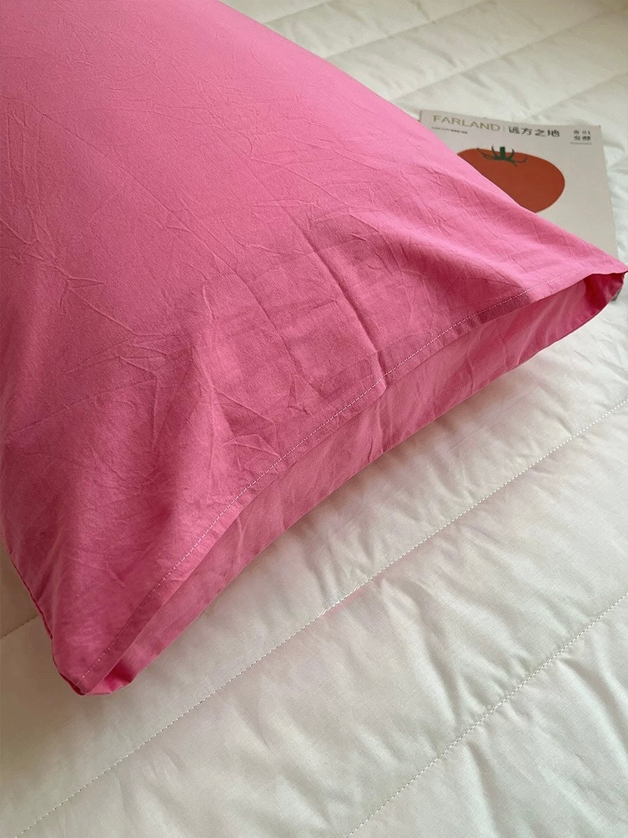A pink pillowcase resting on a white quilted bedspread, with a small book or card partially visible underneath.