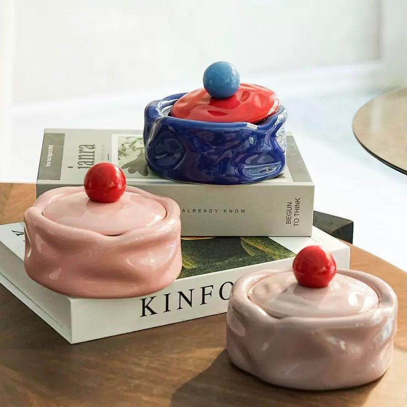 Three ceramic containers with lids on top of books, one pink, one blue, and one white.