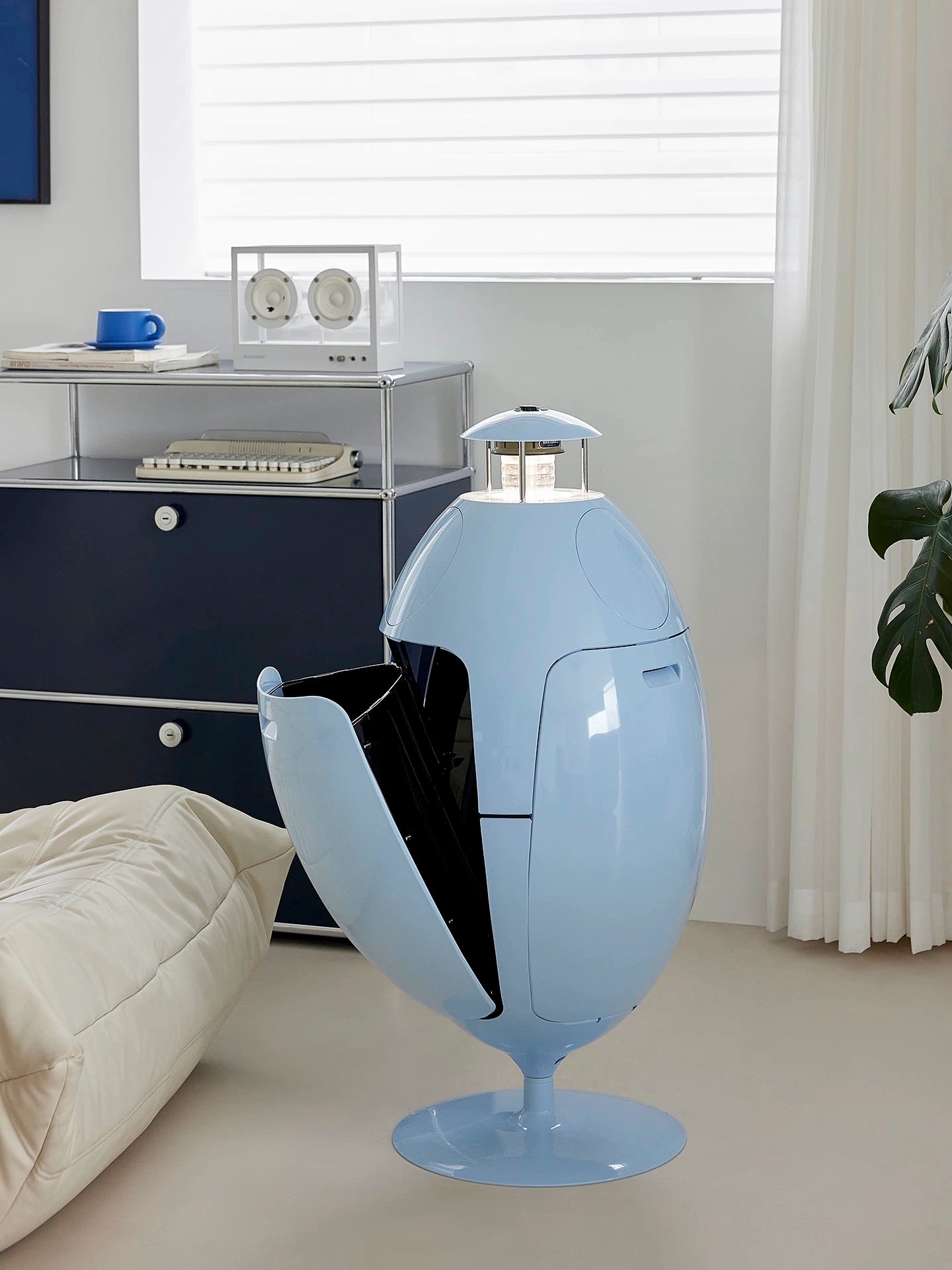 Modern blue trash can in a room with a desk and window blinds.