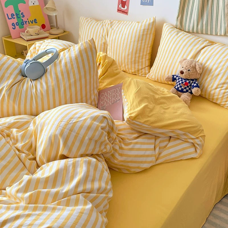 Yellow and white striped bedding set on a bed with a teddy bear and decorative pillows.