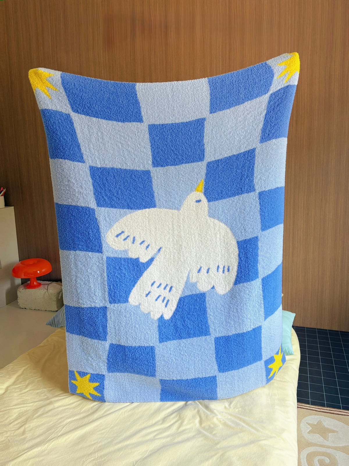 A blue and white checkered blanket with a white dove in the center and yellow star accents, draped over a bed in a bedroom setting.