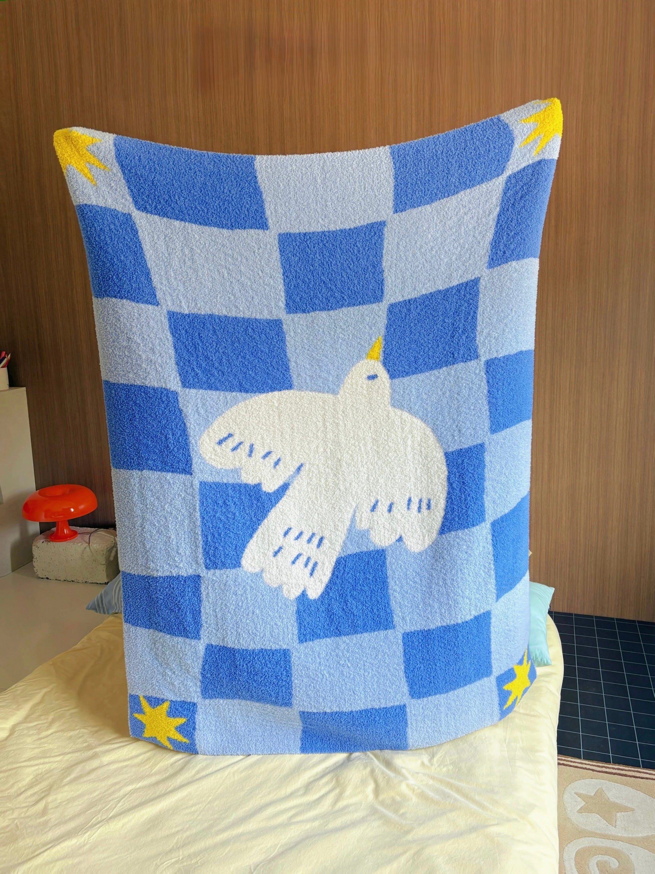 A blue and white checkered blanket with a white dove in the center and yellow star accents, draped over a bed in a bedroom setting.
