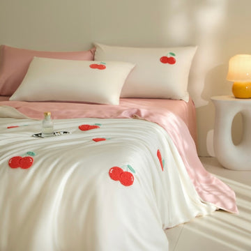 A neatly made bed in a softly lit bedroom, featuring white bedding with embroidered red cherries and a pink satin sheet and pillowcases. A small bedside table with a glowing lamp is visible on the right.