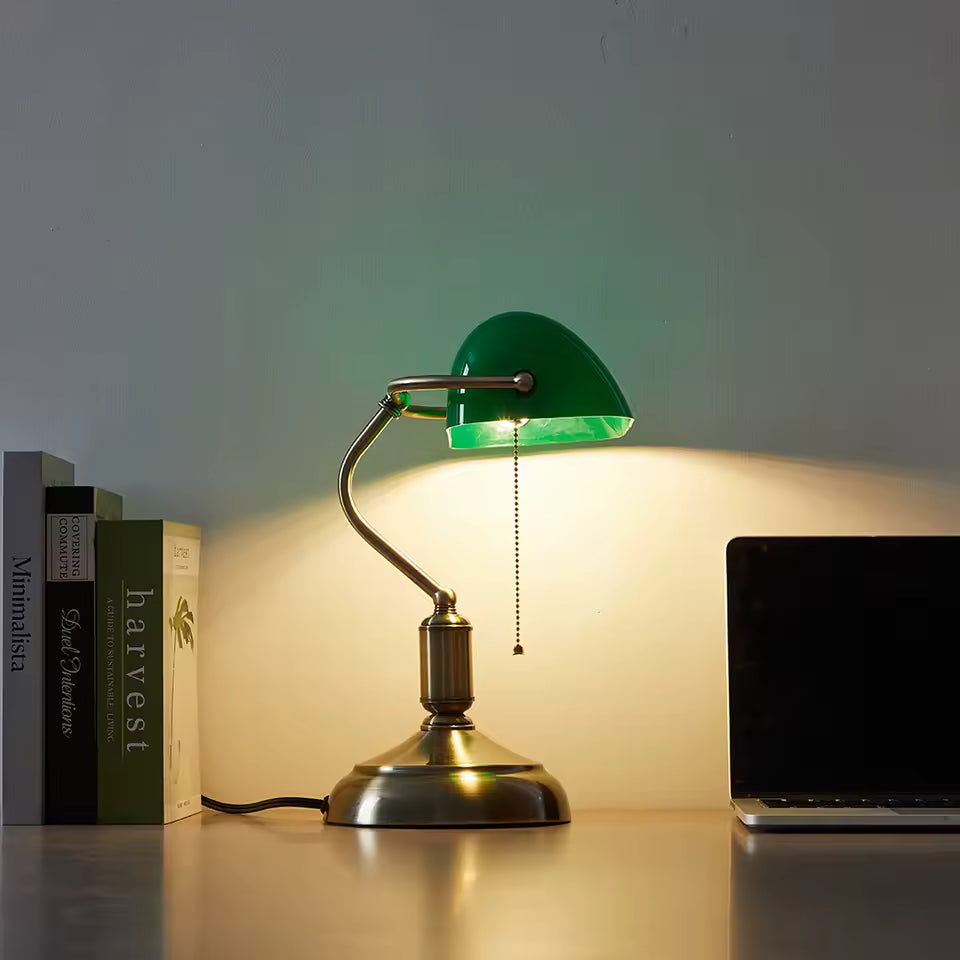 Green desk lamp on a table with books and a laptop in the background