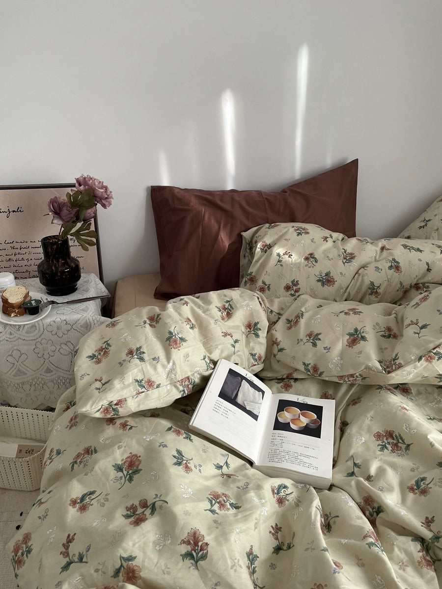 Floral bedding set styled with rust pillows, flowers, open book, and a lace-covered bedside table.