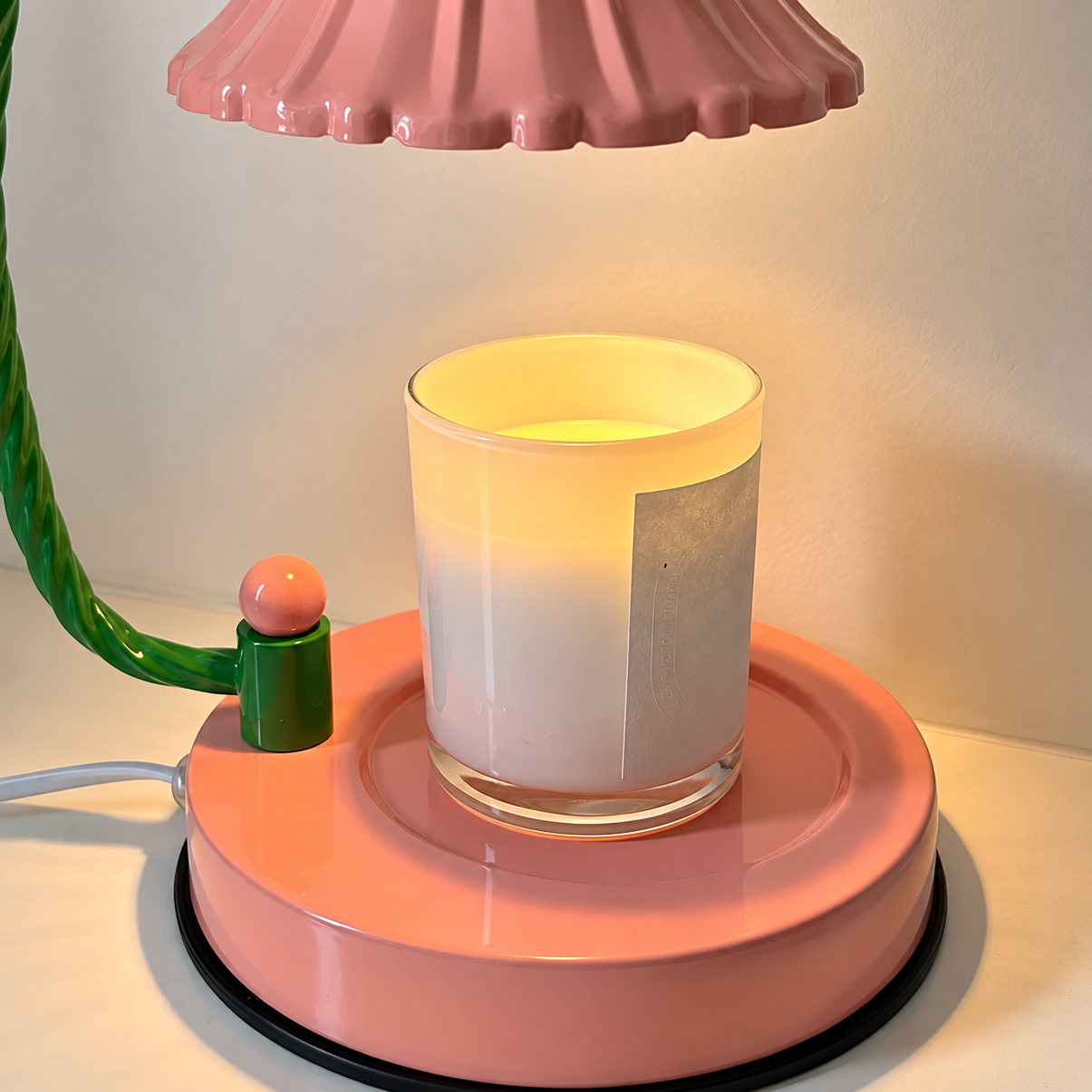 A pink and green table lamp with a scalloped pink shade and a glowing candle placed on its circular base, set against a plain light background.