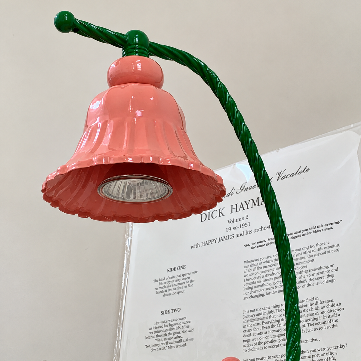 A vintage-style novelty desk lamp with a coral pink bell-shaped shade and a green twisted arm, shown in front of printed sheet music.