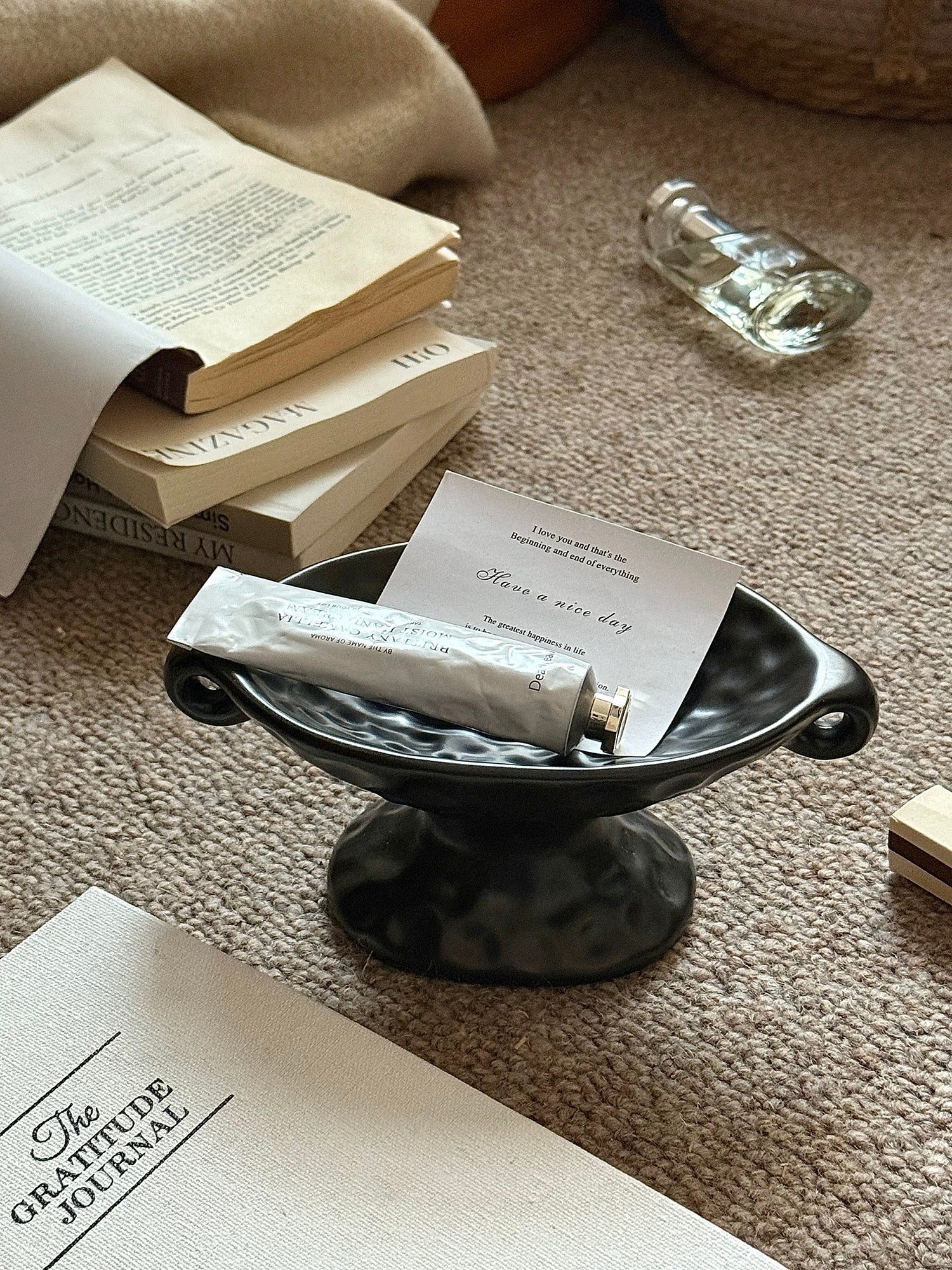A small black ceramic bowl with side handles sits on a beige carpet, holding a white tube and a card. Surrounding props include stacked books, a glass bottle, and a notebook in a styled indoor setting.