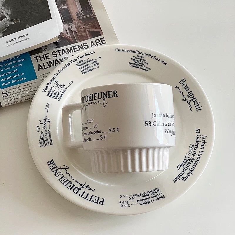 A white ceramic coffee mug with a ribbed handle and base, printed with black French text, resting on a matching saucer with the same text design. The set is styled on a white surface with a magazine in the background.