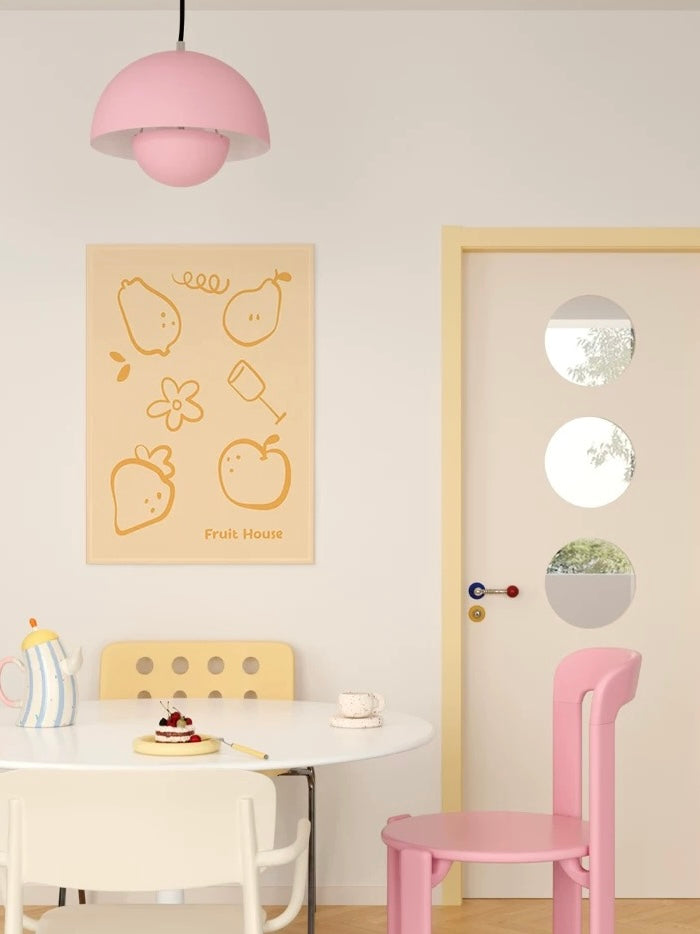 A yellow fruit wall hanging is shown beside a pale door, with a small table and pink chair below.