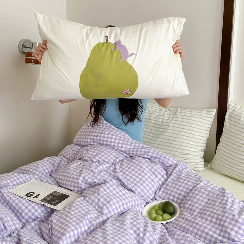 Person holding a pillow with a pear design over their face, lying on a bed with purple checkered duvet and green apples.