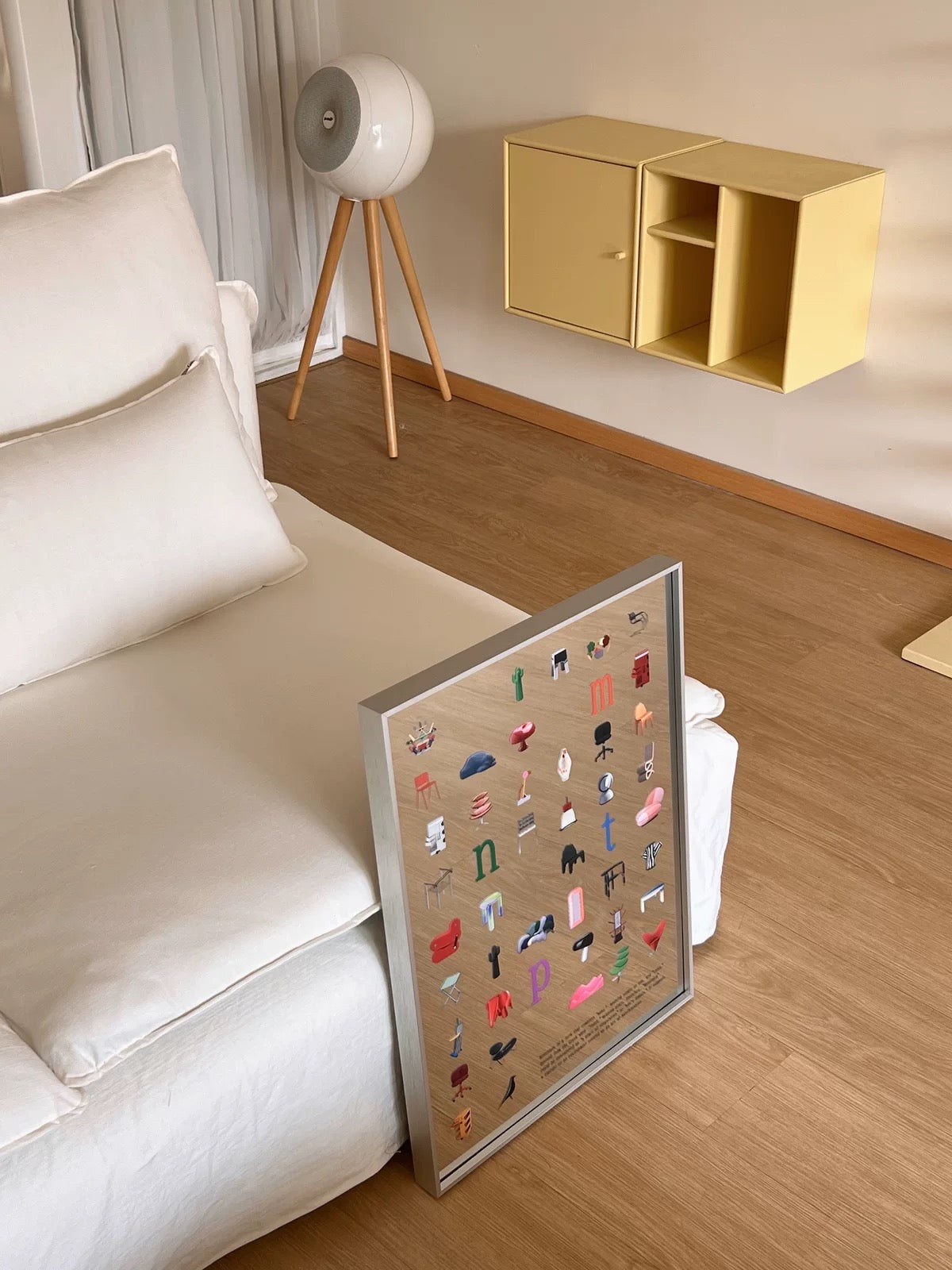 A framed art print featuring a colorful collection of shoe illustrations on a light wood background, leaning against a white sofa in a modern, sunlit room with wooden floors.