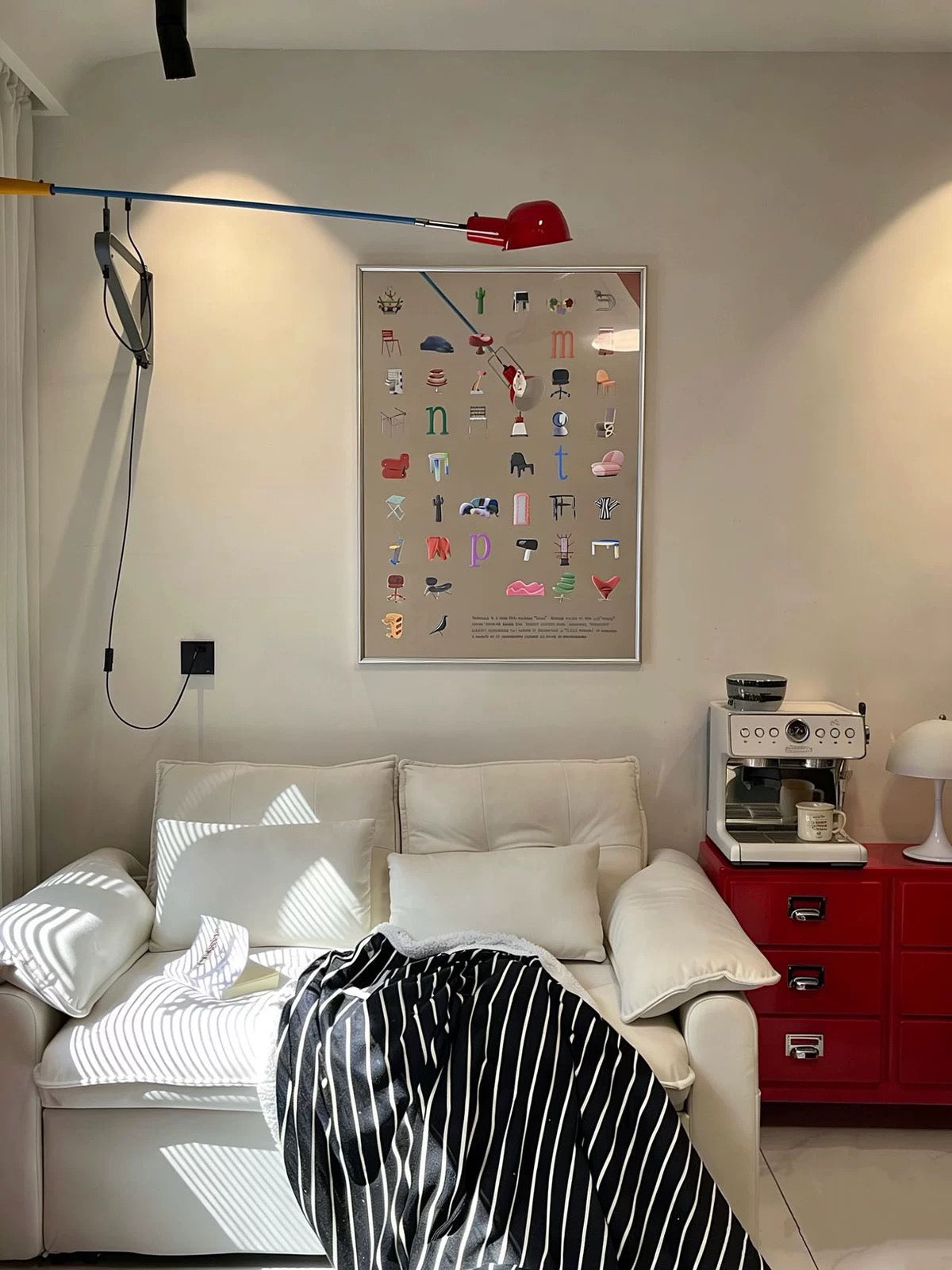 A framed art print featuring a colorful, whimsical clock face design with illustrated objects for each hour, hanging on a beige wall above a white sofa in a modern, stylishly decorated room.