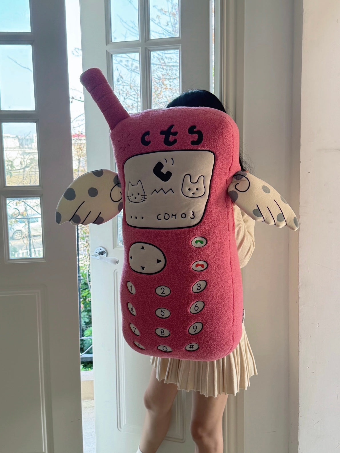 A giant pink phone plush is held upright, showing its long body, number buttons and pale arms with blue spots.