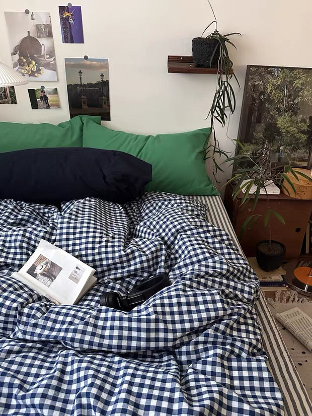 Bed with blue and white checkered blanket, green pillows, and a book on a bed in a room with plants and photos on the wall.