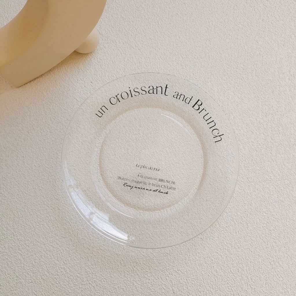 A clear circular glass plate with black cursive text reading 'un croissant and Brunch' on a textured cream surface, with part of a beige ceramic object visible in the top left corner.