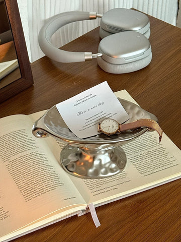 A silver metallic bowl-shaped tray holding an open invitation card and a wristwatch, placed on an open book on a wooden table. A pair of silver over-ear headphones sits in the background.