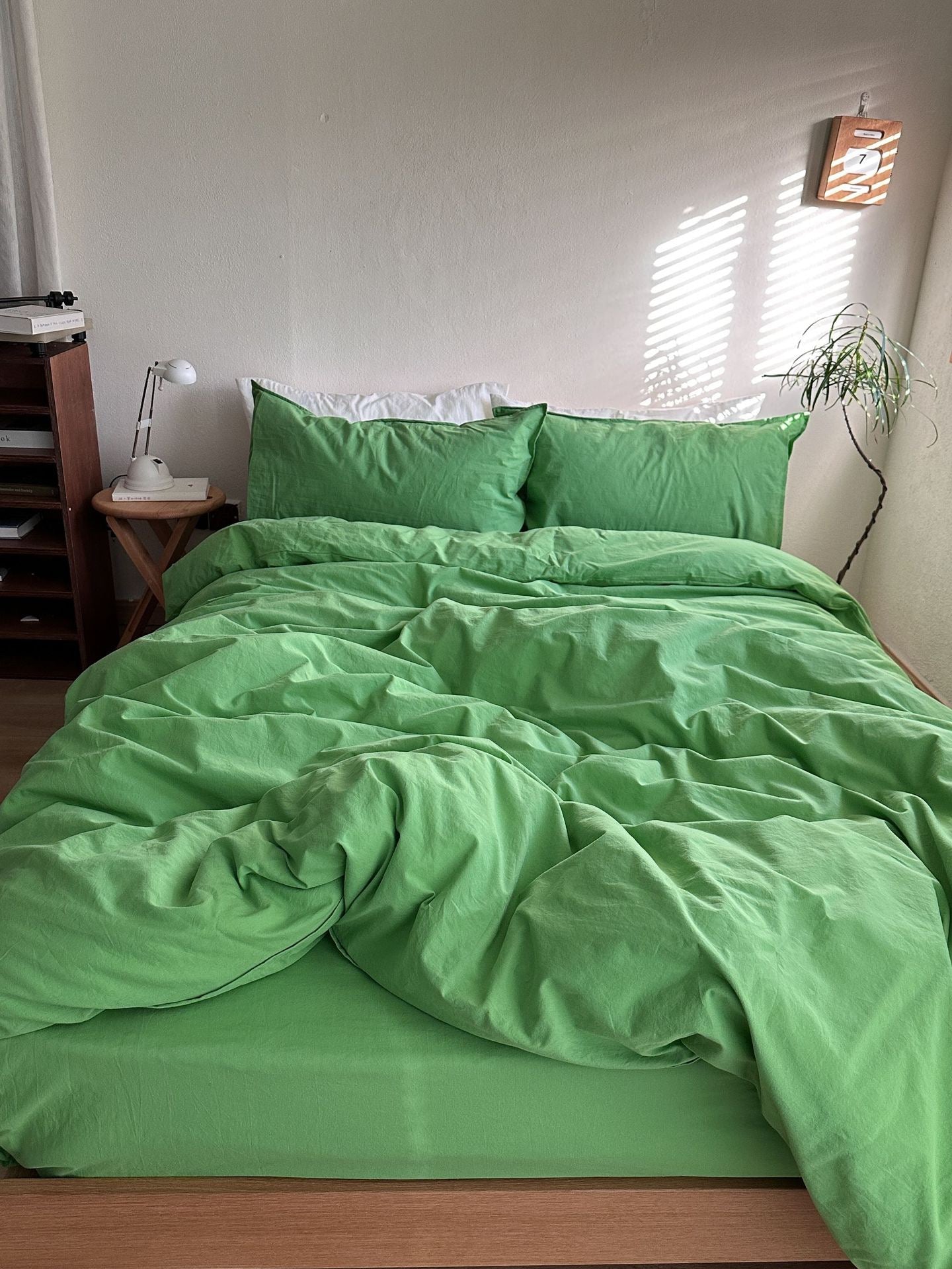 A bed made with a bright green bedding set, including a duvet cover and two matching pillowcases, styled in a minimalist bedroom with a wooden side table, lamp, and plant.