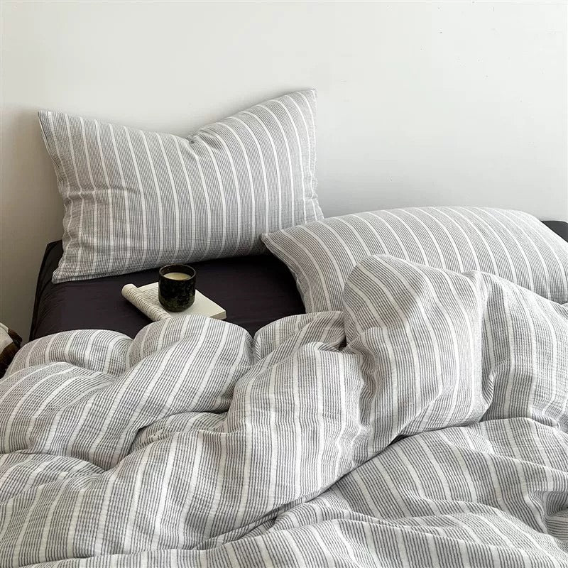 A bed made with a light gray and white striped bedding set, including a duvet cover and two matching pillowcases, styled in a minimalist bedroom setting.