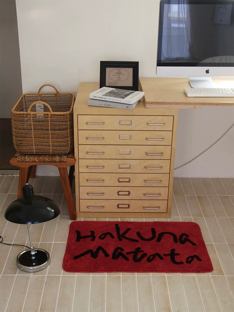 A red Hakuna Matata mat sits in front of a drawer cabinet and next to a black stool on the floor.