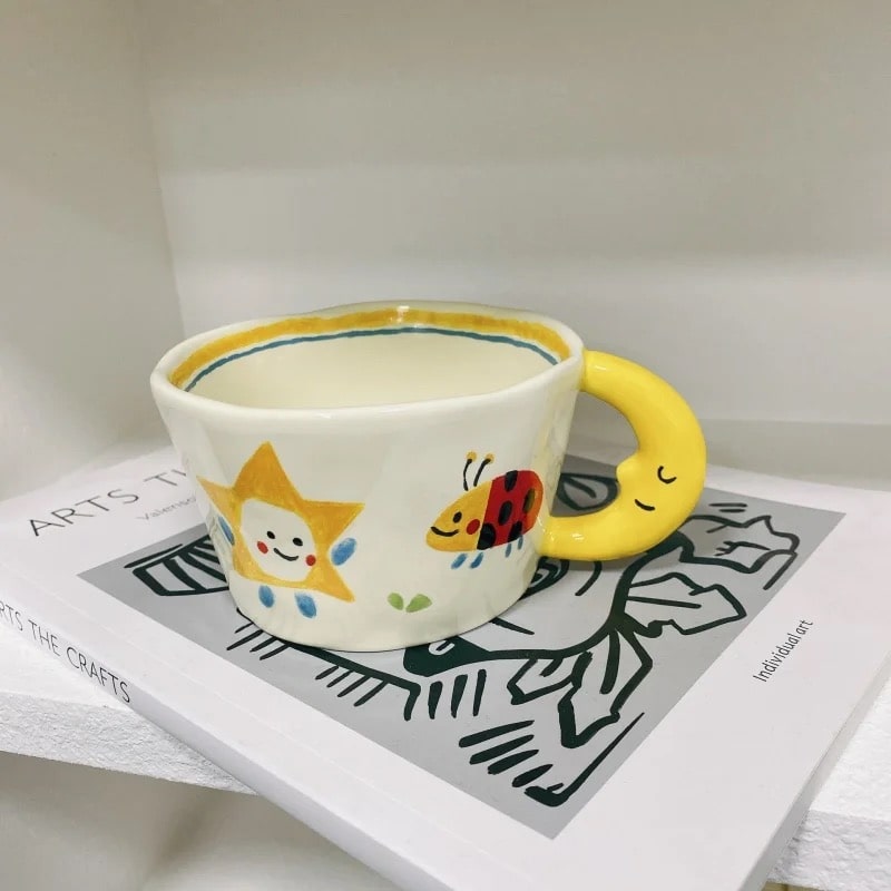 Yellow coffee mug with moon handle and cartoon patterns. Durable ceramic cup for kids, couples, and artistic decor.